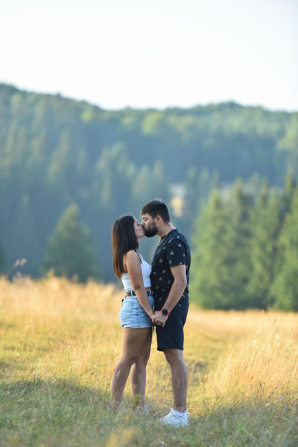 Couple sharing intimate moments in a scenic meadow with evergreen forest backdrop.