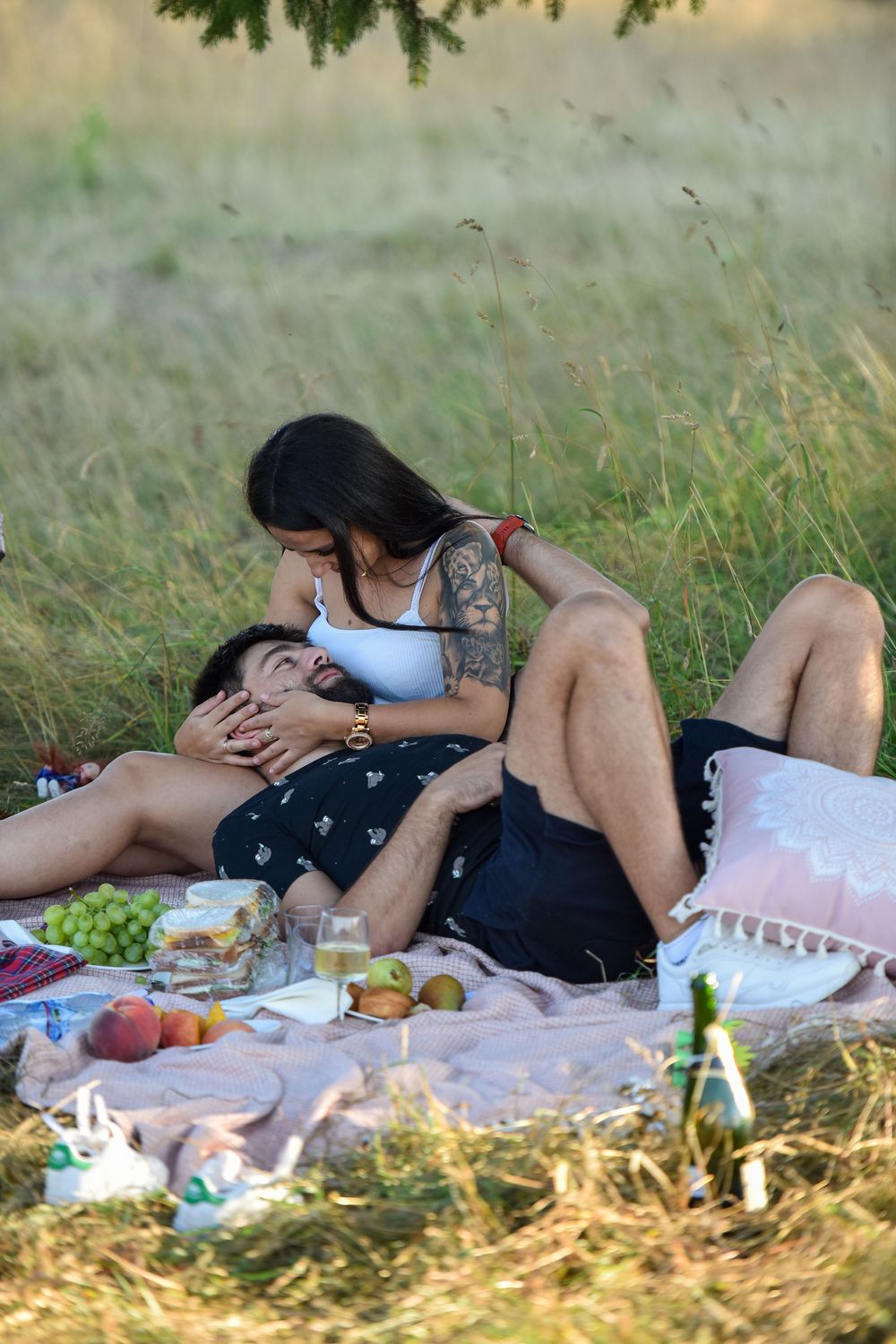 A couple relaxes on a picnic blanket in a grassy field with food and drinks on a sunny summer day.