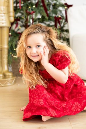 A child in a bright red holiday dress twirls and plays on a wooden floor near Christmas decorations.
