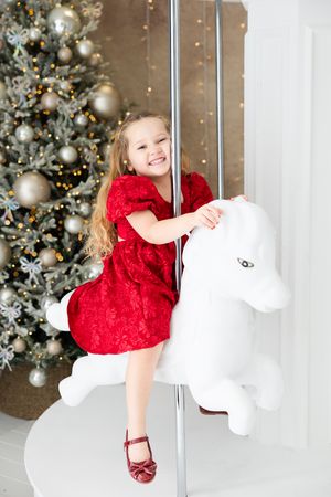 A child in a festive red dress plays with white decorations near a Christmas tree with silver ornaments.