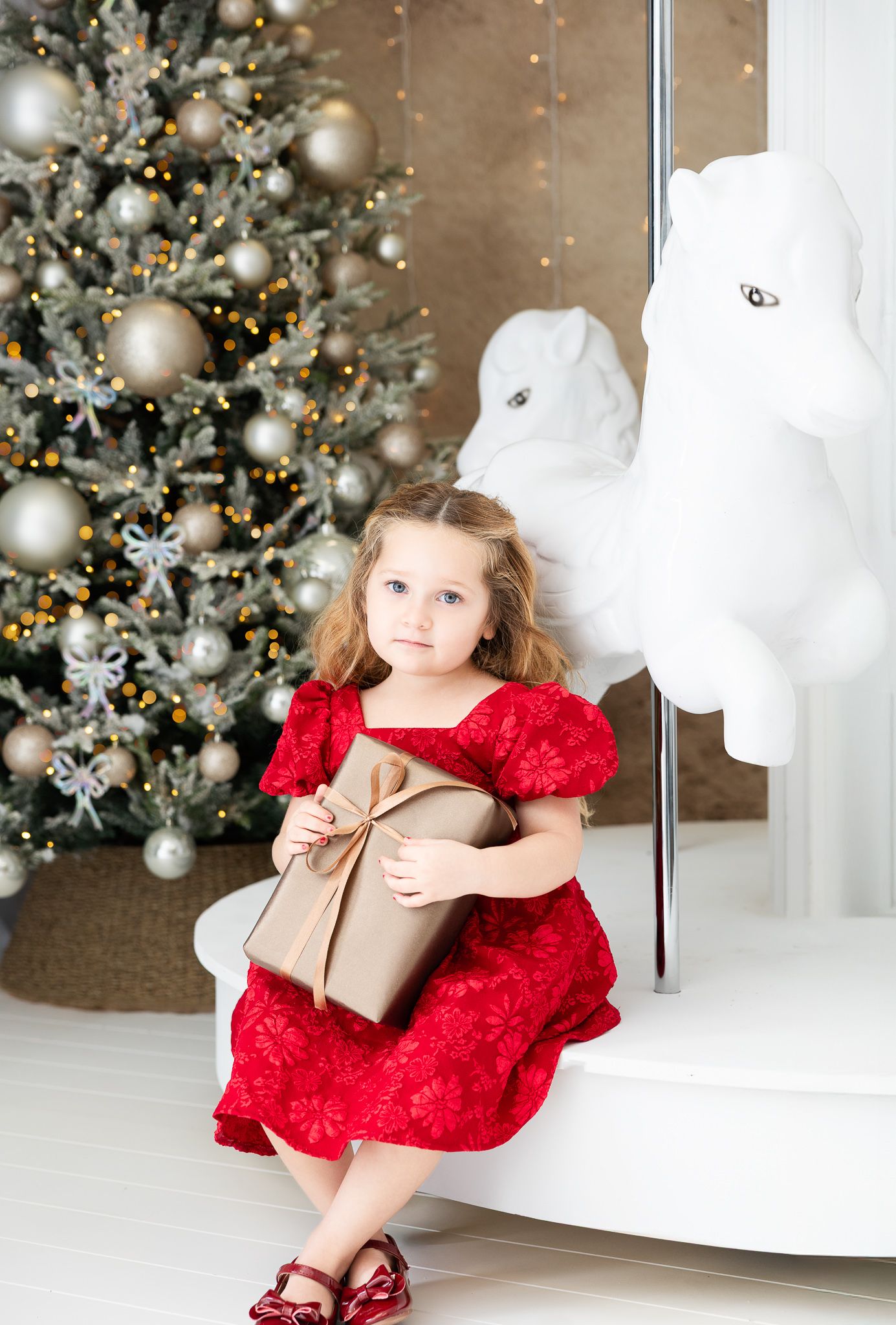 A young child in a red holiday dress sits by a decorated Christmas tree holding a wrapped gift.