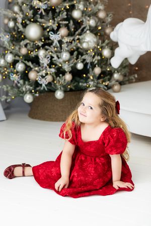 A child in a red velvet holiday dress poses on a white floor with decorated Christmas trees and ornaments in the background.