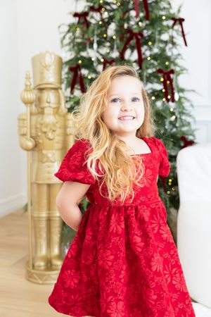 A child in a red velvet holiday dress twirls and plays in front of a decorated Christmas tree and gold nutcrackers.