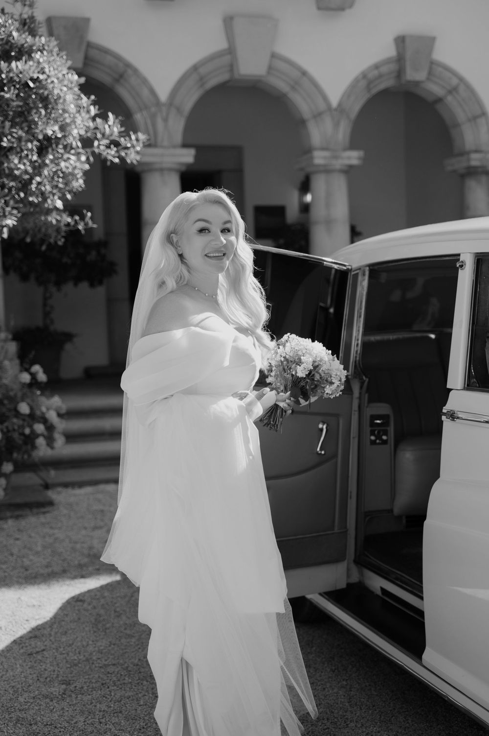 A bride in a flowing white dress enters a classic white vintage car in a sequence of black and white photos.