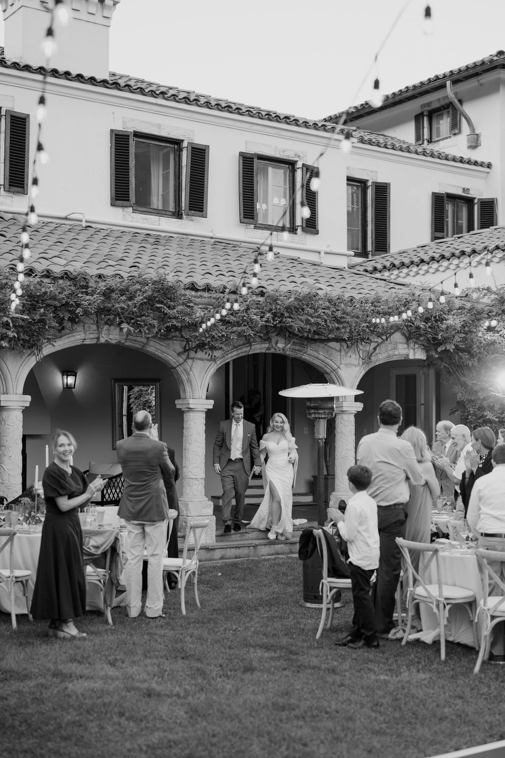 Black and white photo of an outdoor garden party with guests mingling under string lights at a Spanish-style villa.