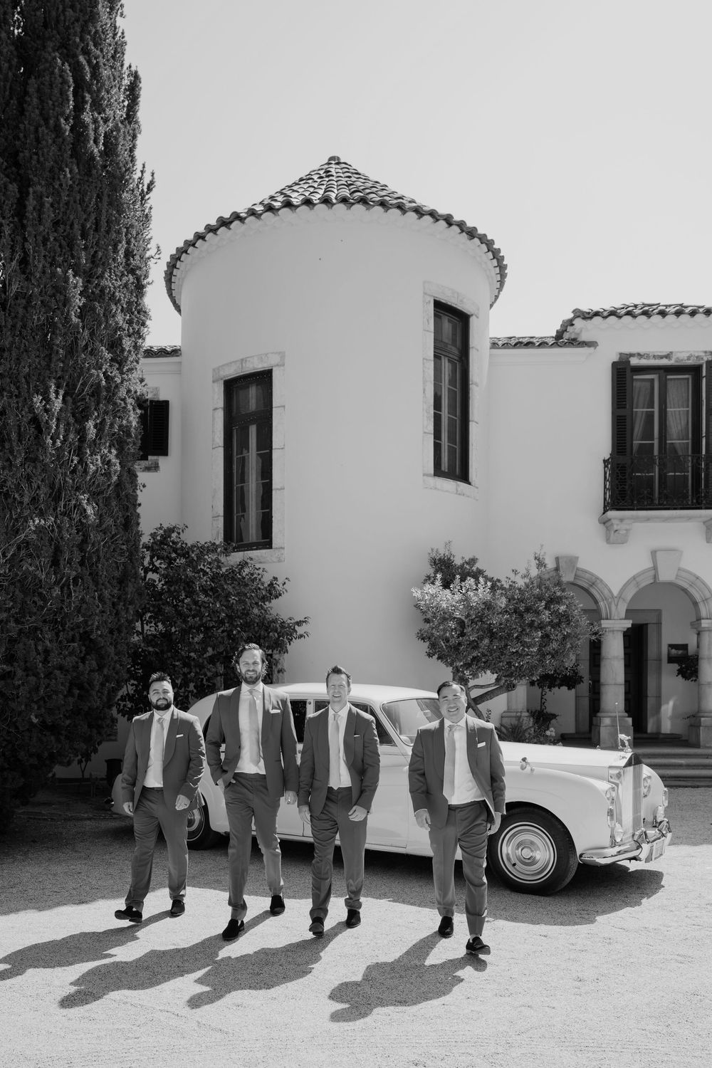 A black and white photo series showing groomsmen posing in formal attire next to a vintage car outside a Spanish-style building.