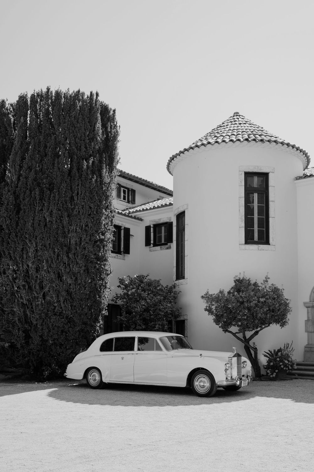 A classic white luxury car parked beside a Mediterranean-style white stucco building with black shutters and tall cypress trees.