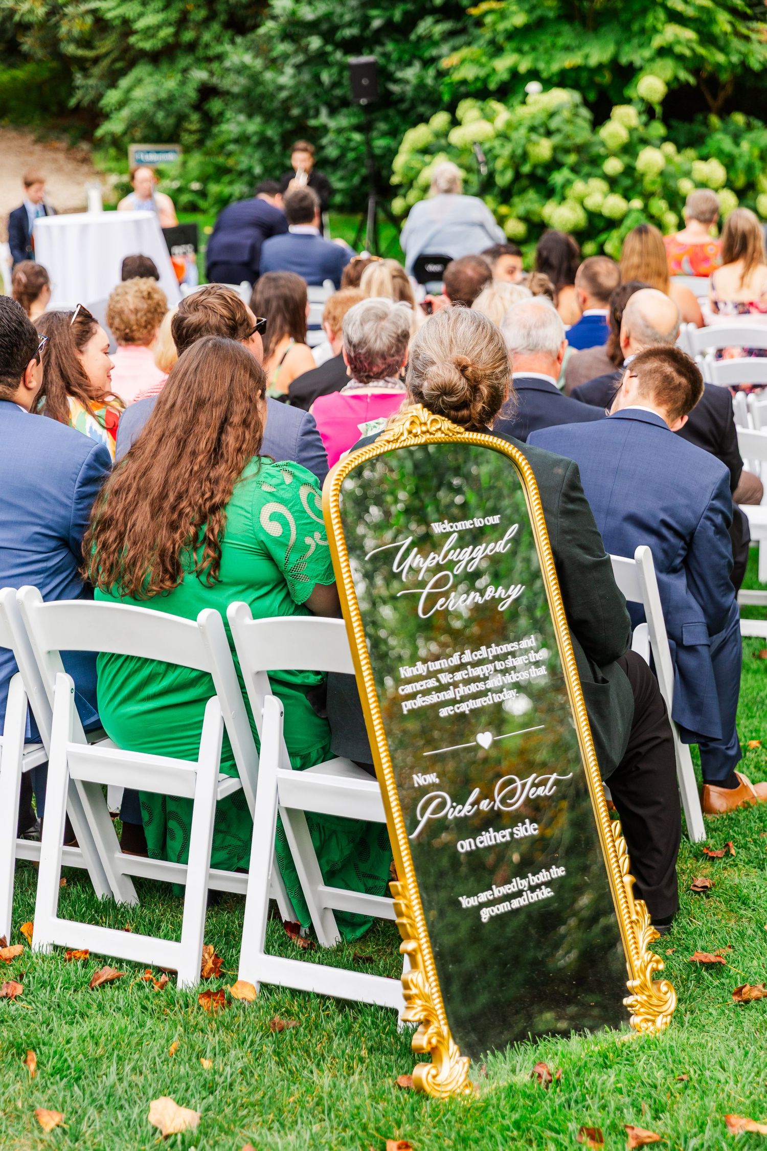 Wedding guests seated in white chairs with green decor during an outdoor garden ceremony.