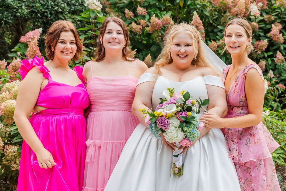 A group in pink dresses and a white wedding gown pose together in a garden setting for formal photos.