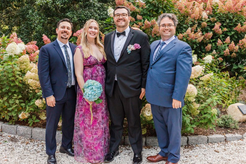 Wedding party poses together outdoors with pink floral backdrop and coordinated navy and pink attire.