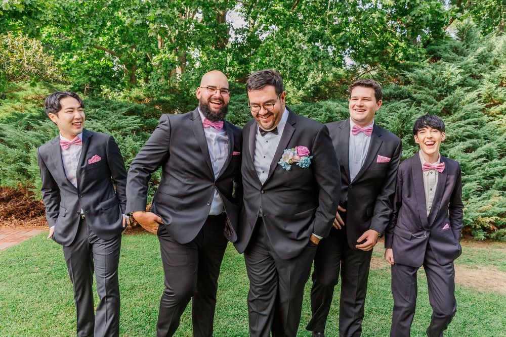 Five groomsmen in formal suits pose together on a garden path surrounded by lush green foliage.