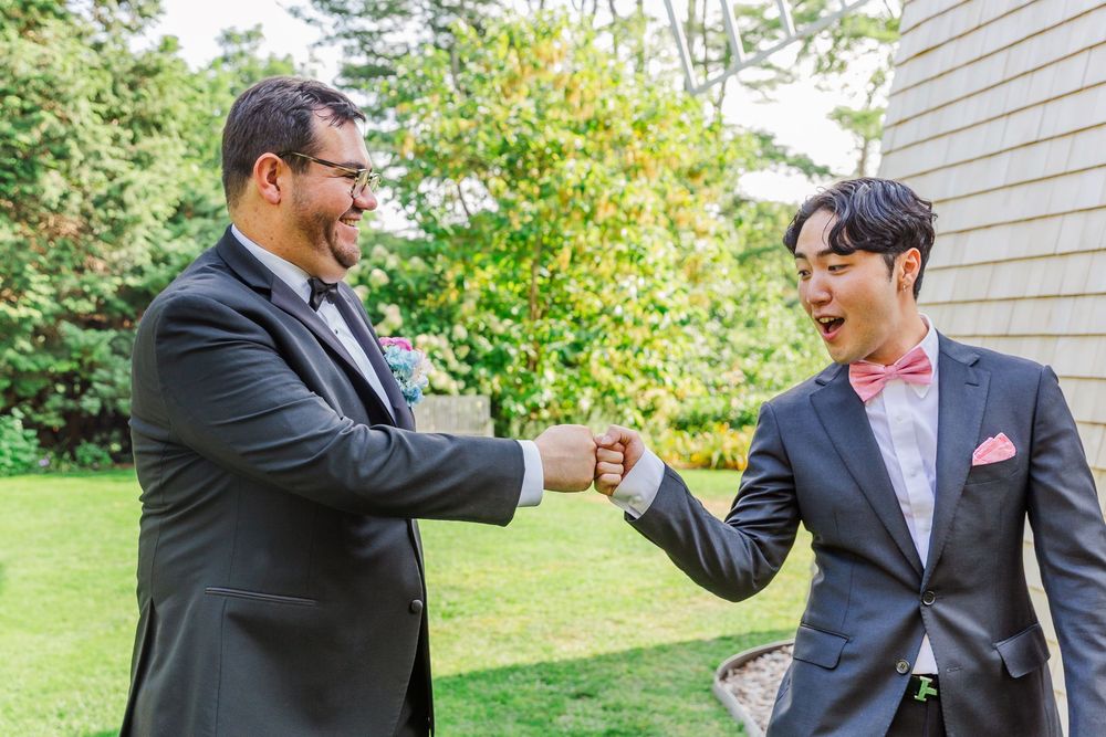 Two men in dark suits share a celebratory fist bump outdoors with trees and siding visible in the background.