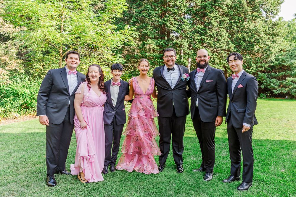 Wedding party in pink dresses and dark suits poses together on green lawn during outdoor celebration.