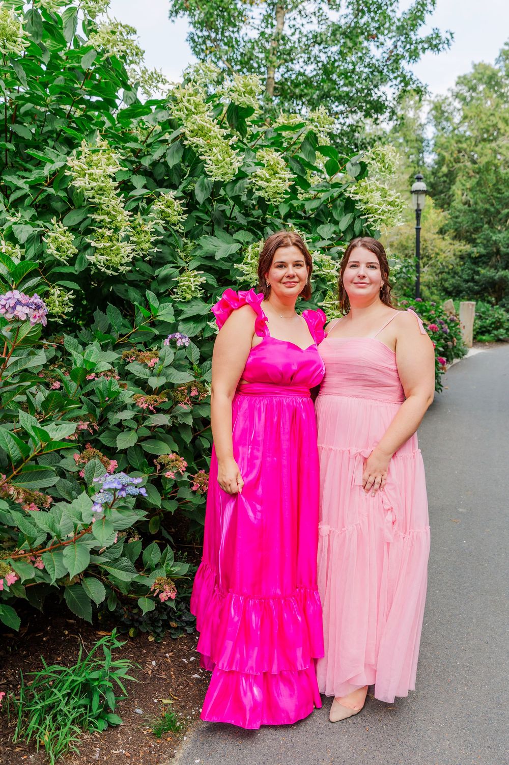 Two people in pink and hot pink formal dresses pose together on a garden path surrounded by lush greenery.