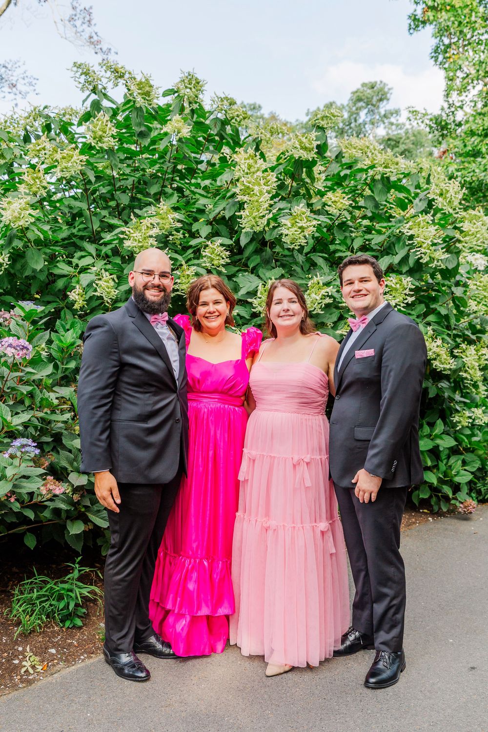 A group in formal attire including black suits and bright pink dresses pose together in a garden setting.