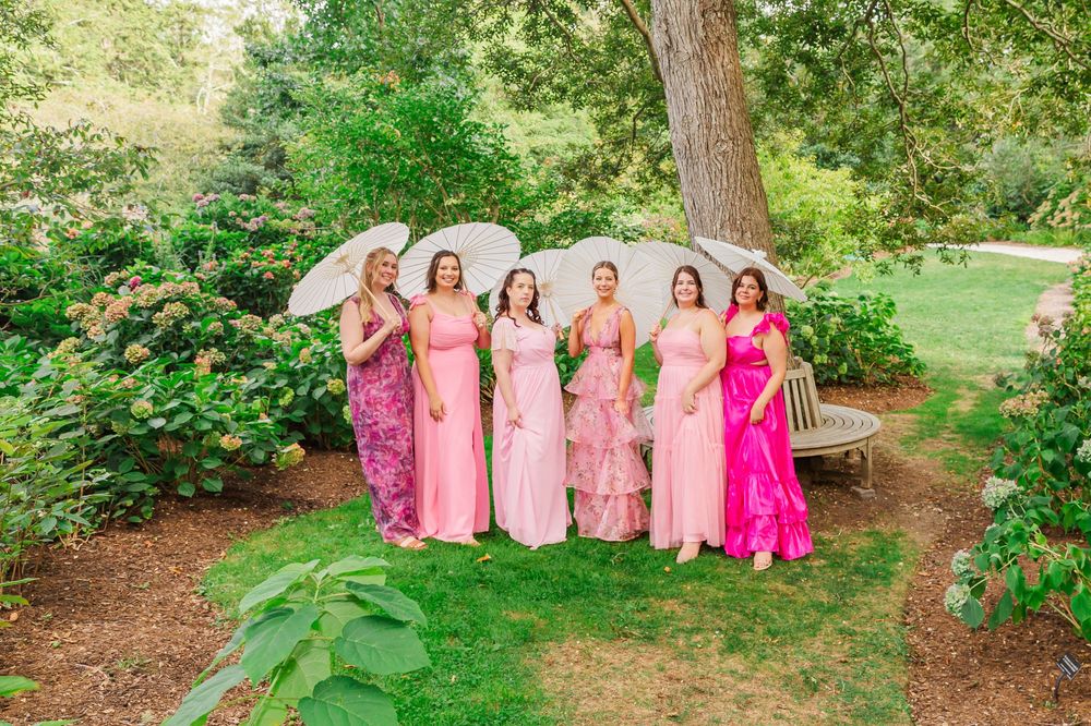 A group in coordinating pink dresses poses together in a lush garden setting for formal photographs.