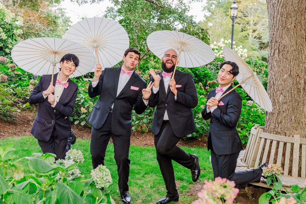 Wedding party members in black suits pose playfully with white parasols in a garden setting.