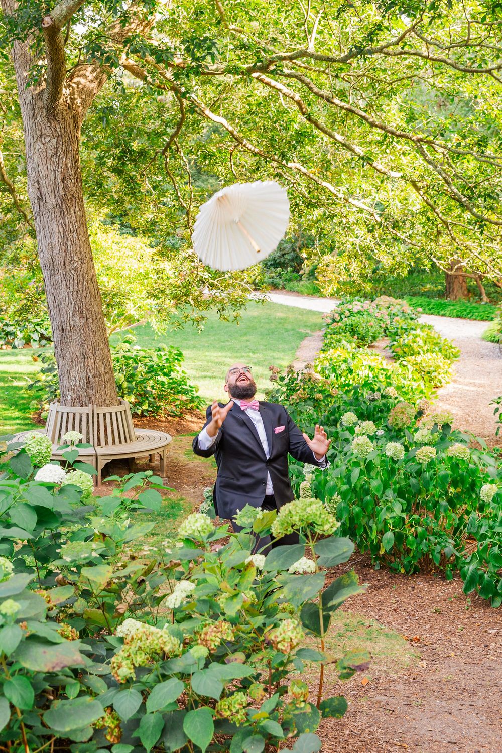 Wedding party members stand with decorative parasols in a lush garden setting with trees and greenery.