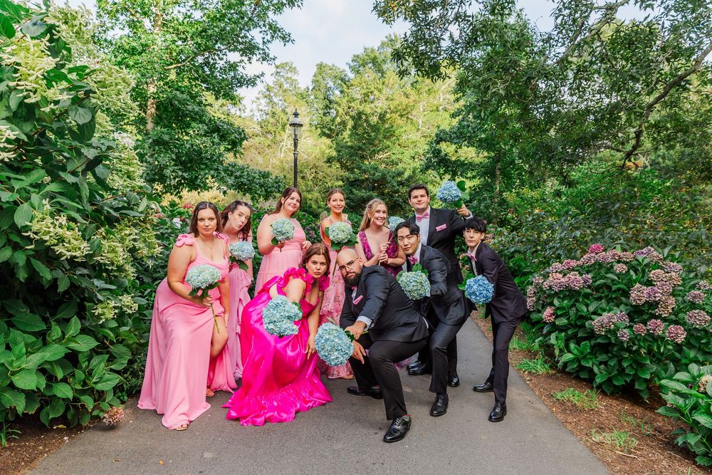 Wedding party members in colorful pink and coral dresses walk along a lush garden path during outdoor celebration.