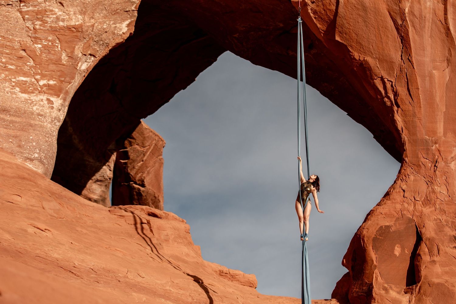 Aerialist suspended on silks under a Utah red rock arch; refined aerial arts photography capturing scale, texture, and motion.