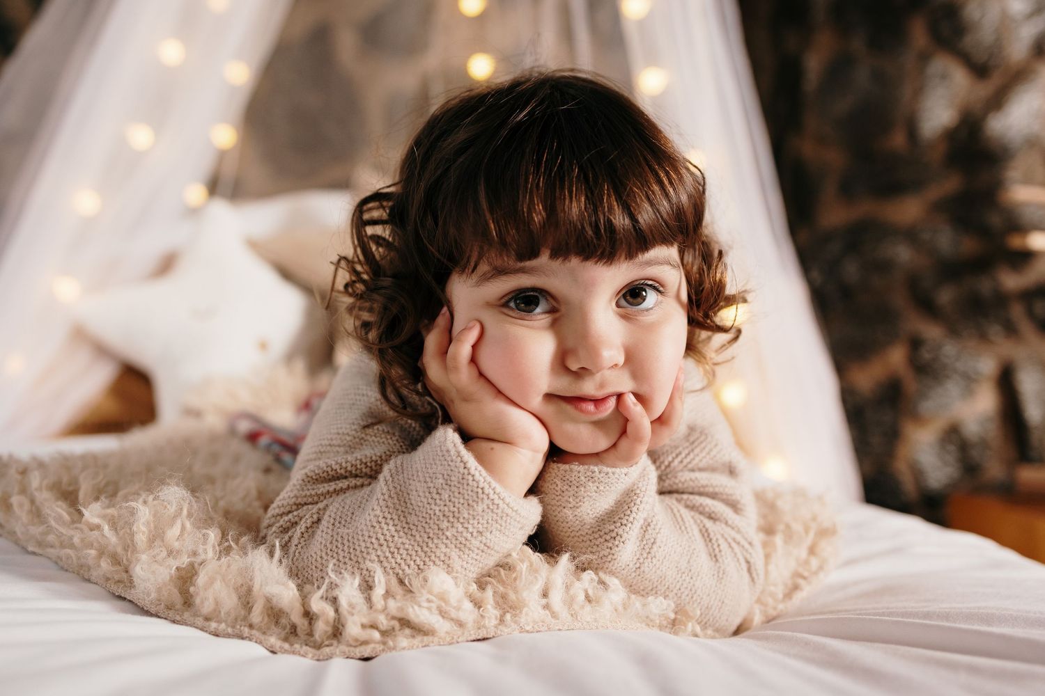 Child laying on cream colored bedding with twinkling lights and sheer curtains in background.