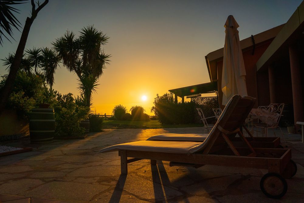 Wooden lounge chair on patio with palm trees silhouetted against golden sunset.