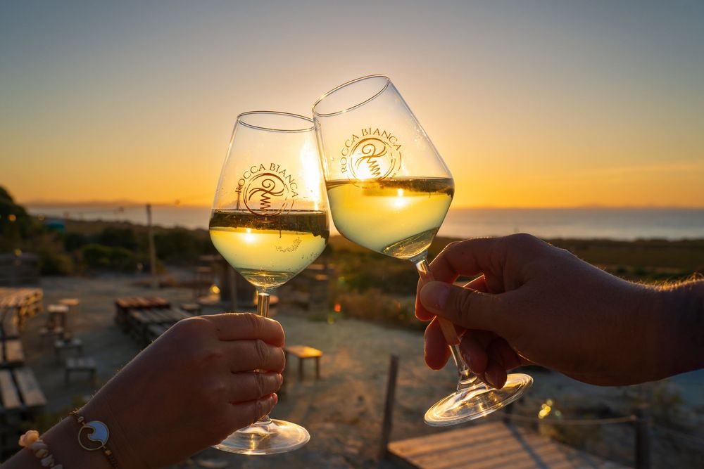 Wine glasses toasting against a coastal sunset with golden light reflecting in the white wine.