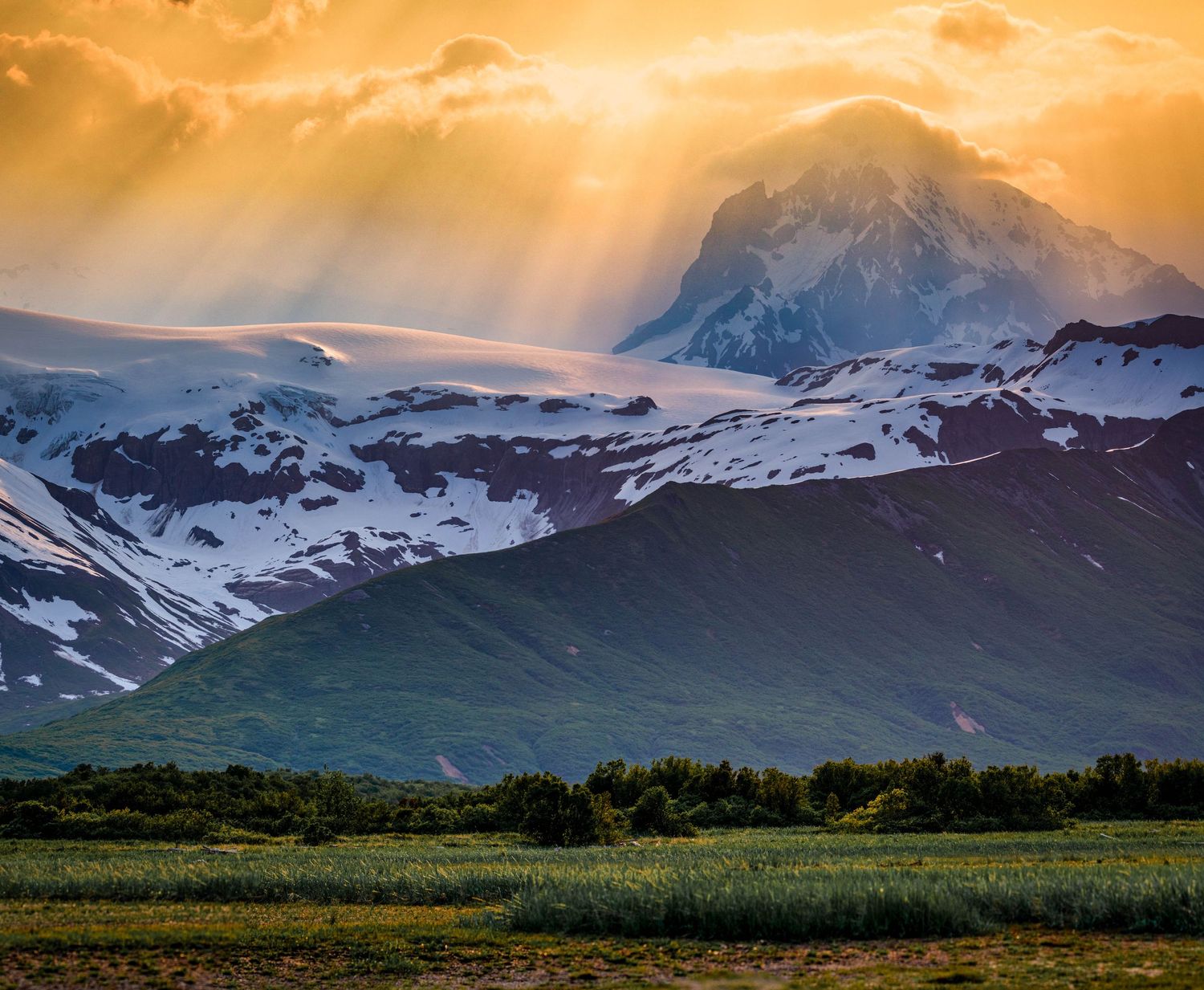Dramatic sunset rays illuminate snowy mountain peaks above green meadows in a majestic alpine landscape.