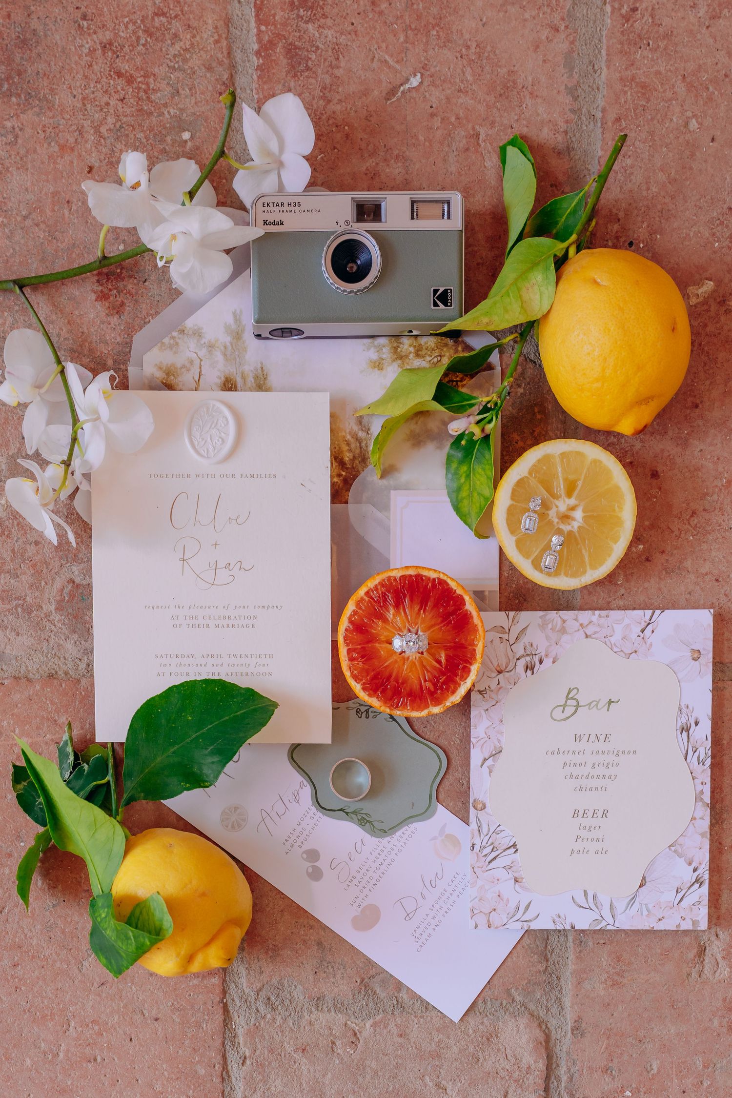 Wedding stationery flatlay with citrus fruits and elegant calligraphy on coral background.