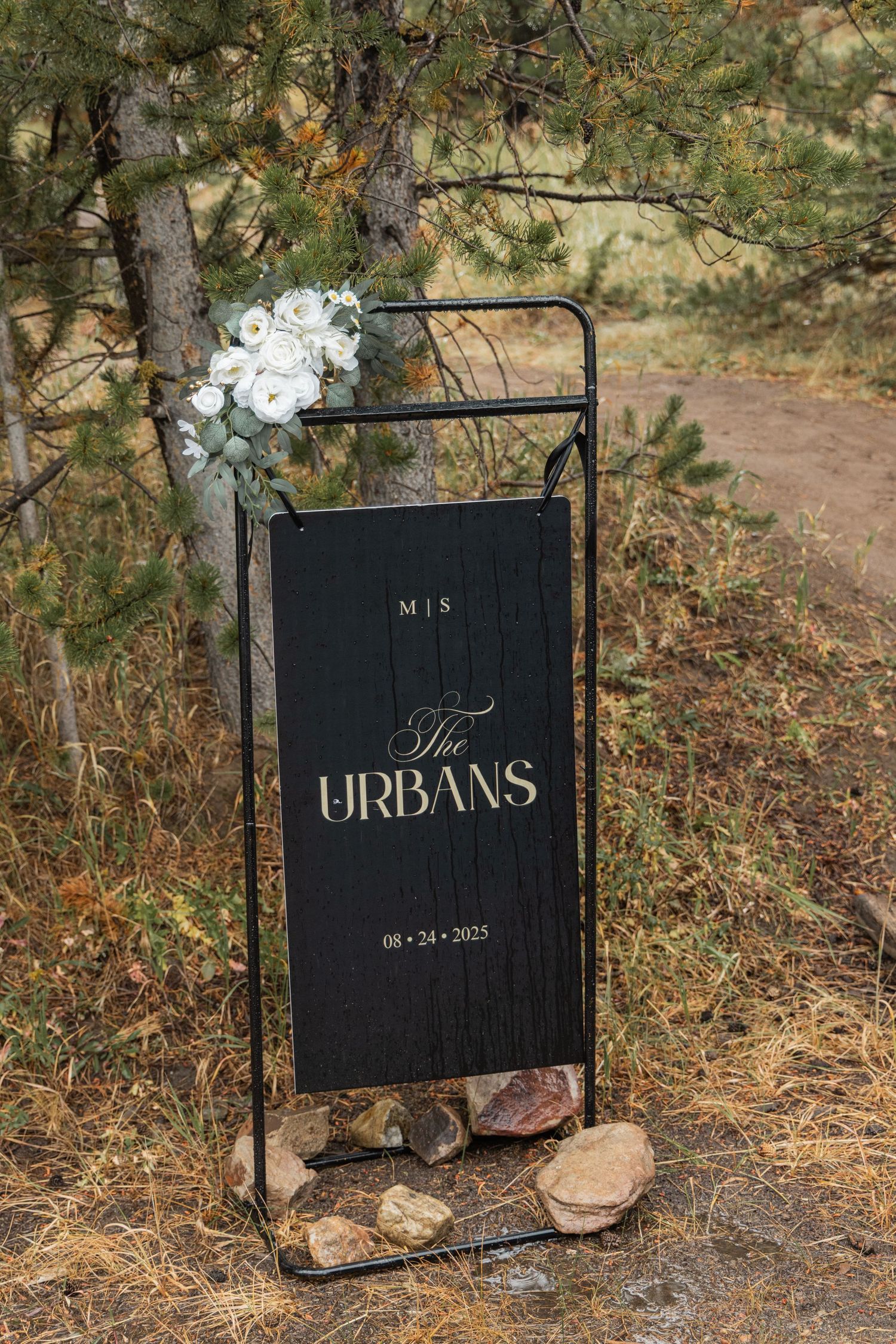 Black wedding sign with 'URBANS' text displayed on ground among pine trees and rocks in outdoor setting.
