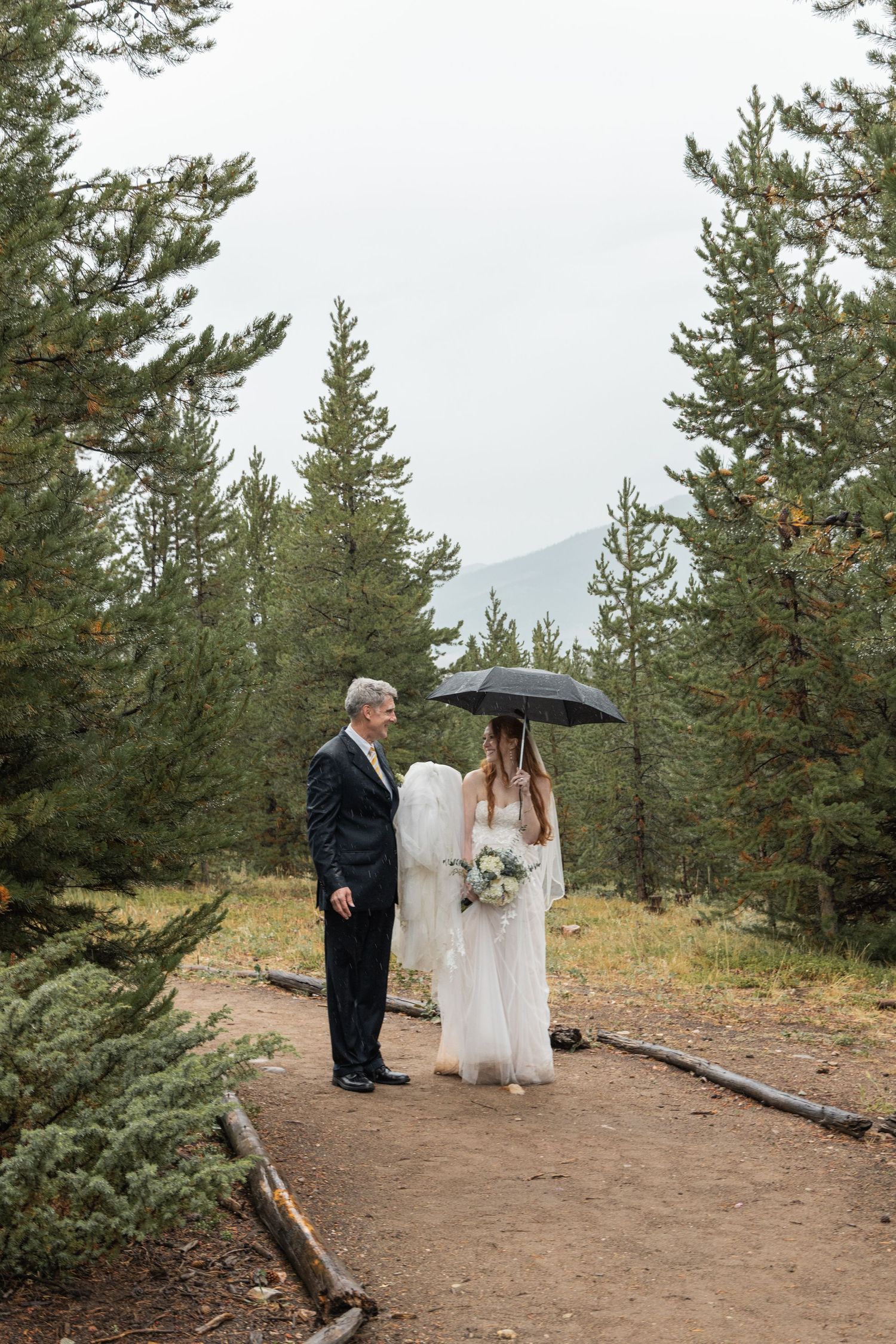 A wedding ceremony takes place on a mountain trail surrounded by evergreen trees on an overcast day.