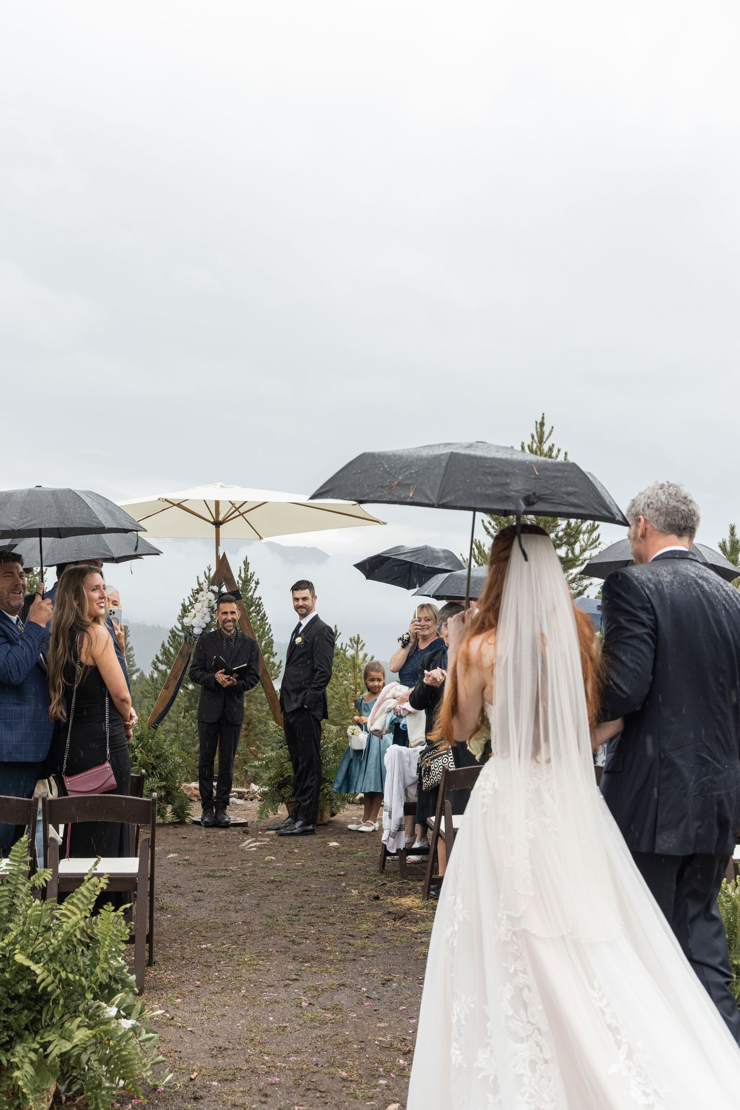 Wedding guests huddle under umbrellas during an outdoor ceremony on a rainy day.