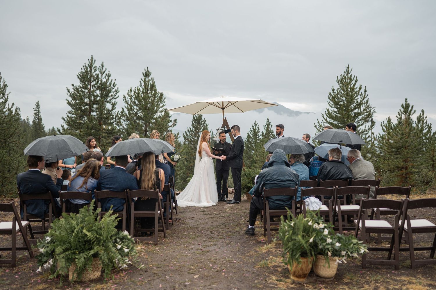 Wedding guests hold umbrellas during an outdoor mountain ceremony on a rainy day.