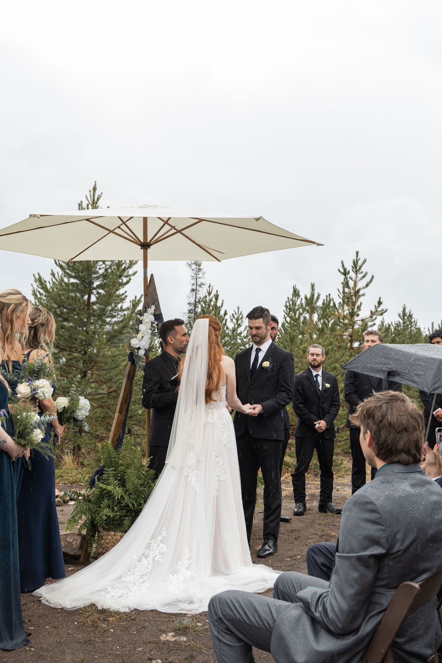An outdoor wedding ceremony takes place under a white umbrella with evergreen trees and guests in navy and grey attire.