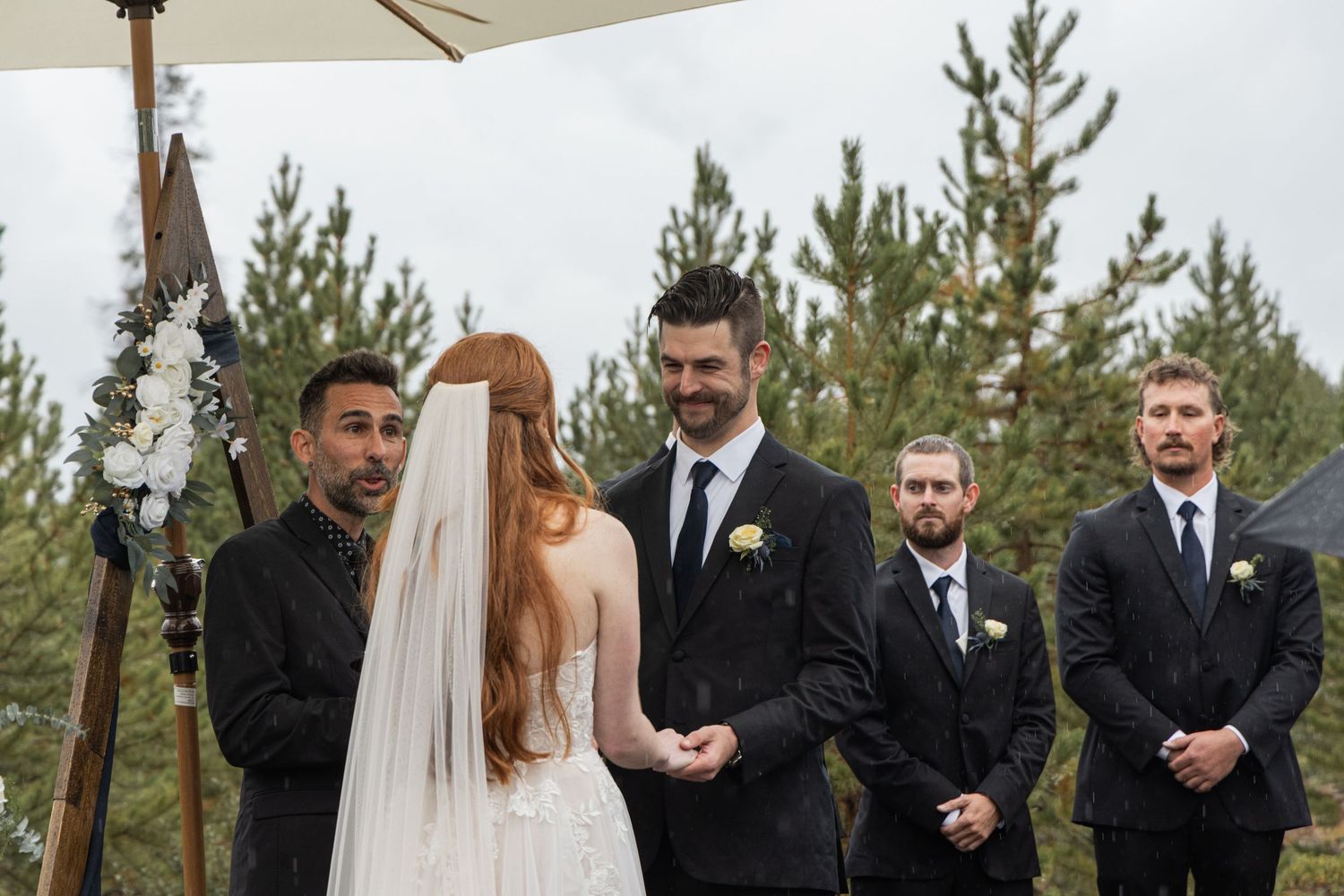 A wedding ceremony takes place outdoors under a wooden arch decorated with white fabric and pine trees in the background.