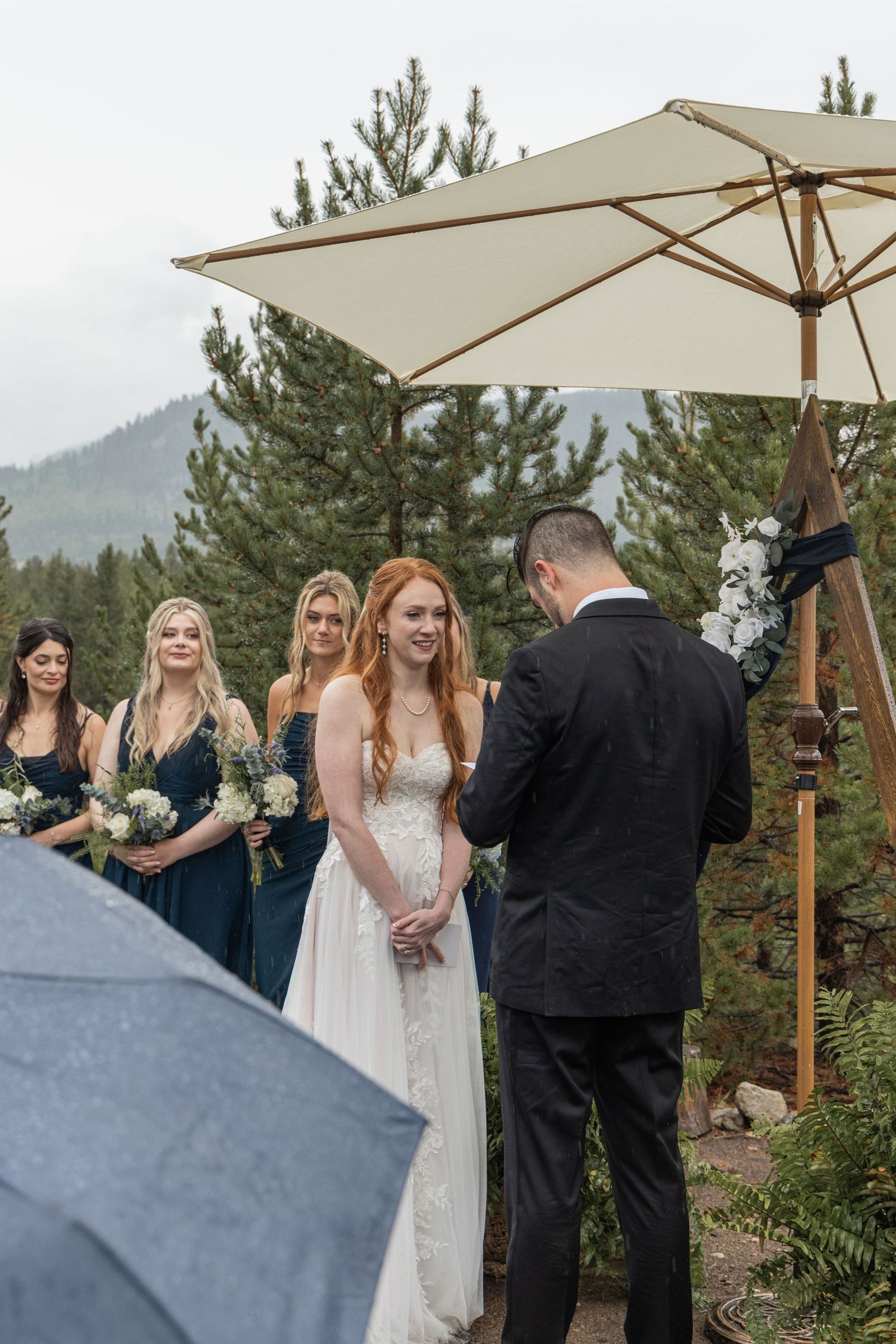 Outdoor wedding ceremony under cream umbrellas with pine trees and mountain backdrop.