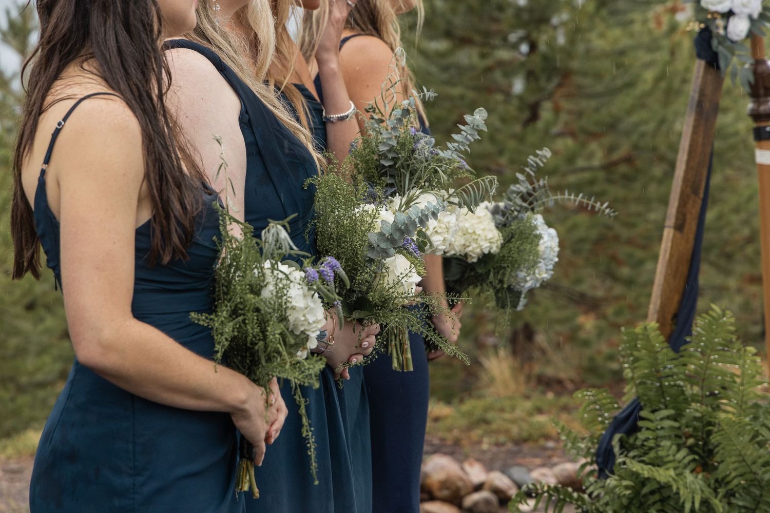 Bridesmaids in navy blue dresses hold white and green floral bouquets during an outdoor wedding ceremony.
