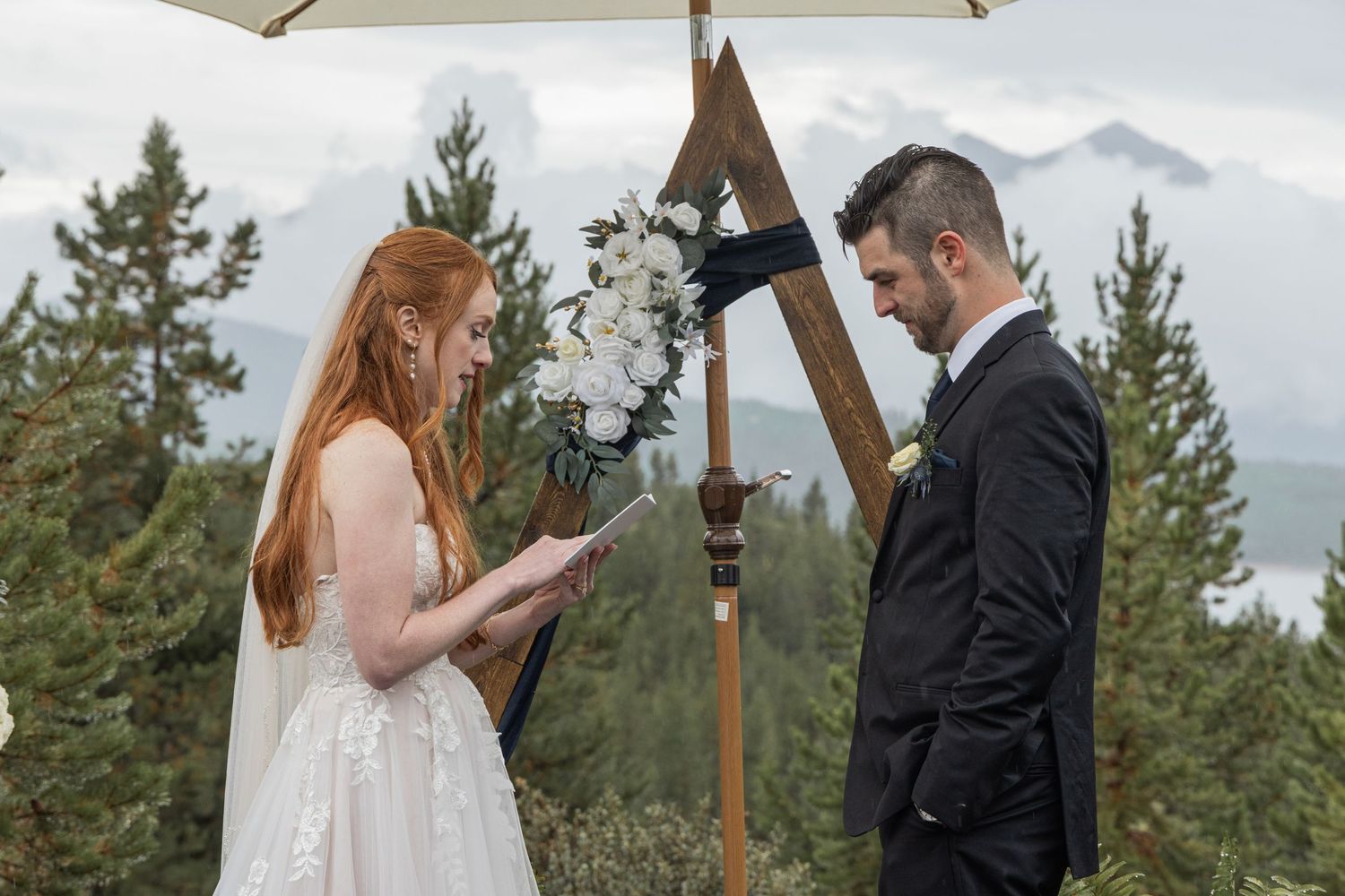 Romantic wedding couple embraces in front of snow-capped mountains and evergreen trees during outdoor ceremony.