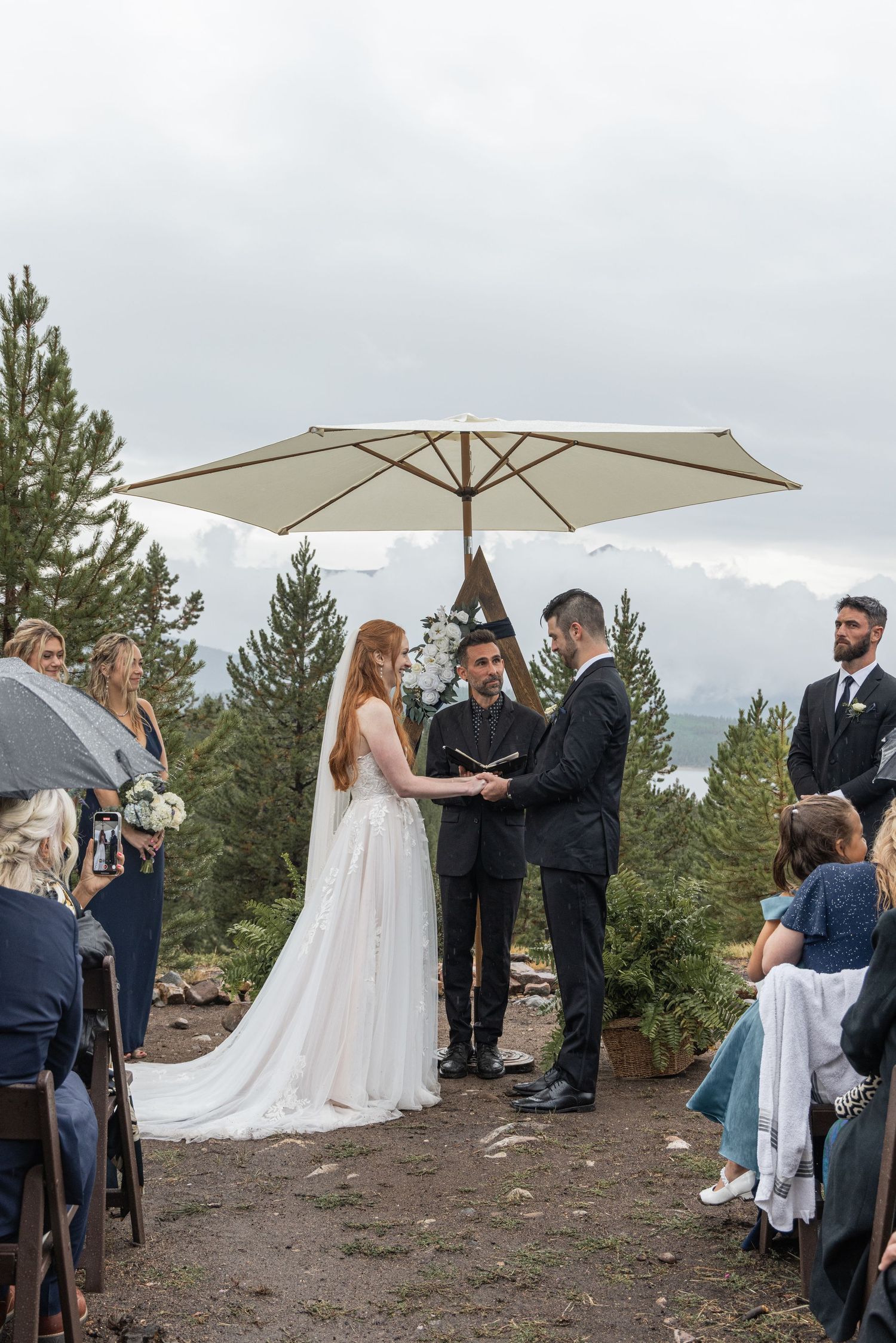 An outdoor wedding ceremony takes place under a white umbrella with evergreen trees and cloudy skies in the background.