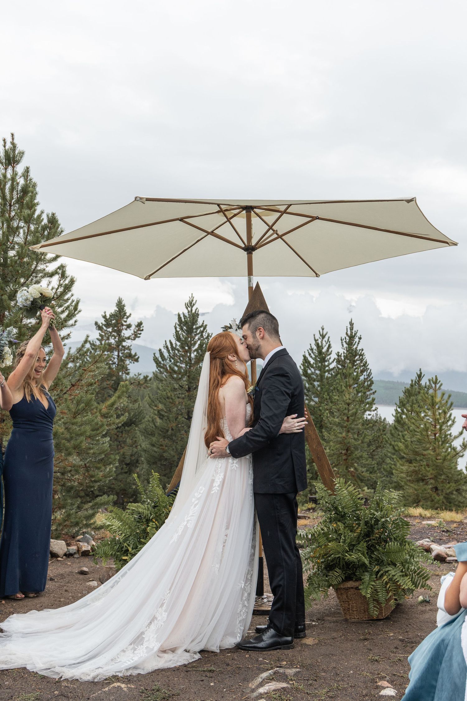 A couple shares a romantic moment under a white umbrella on a cloudy mountaintop with pine trees in the background.