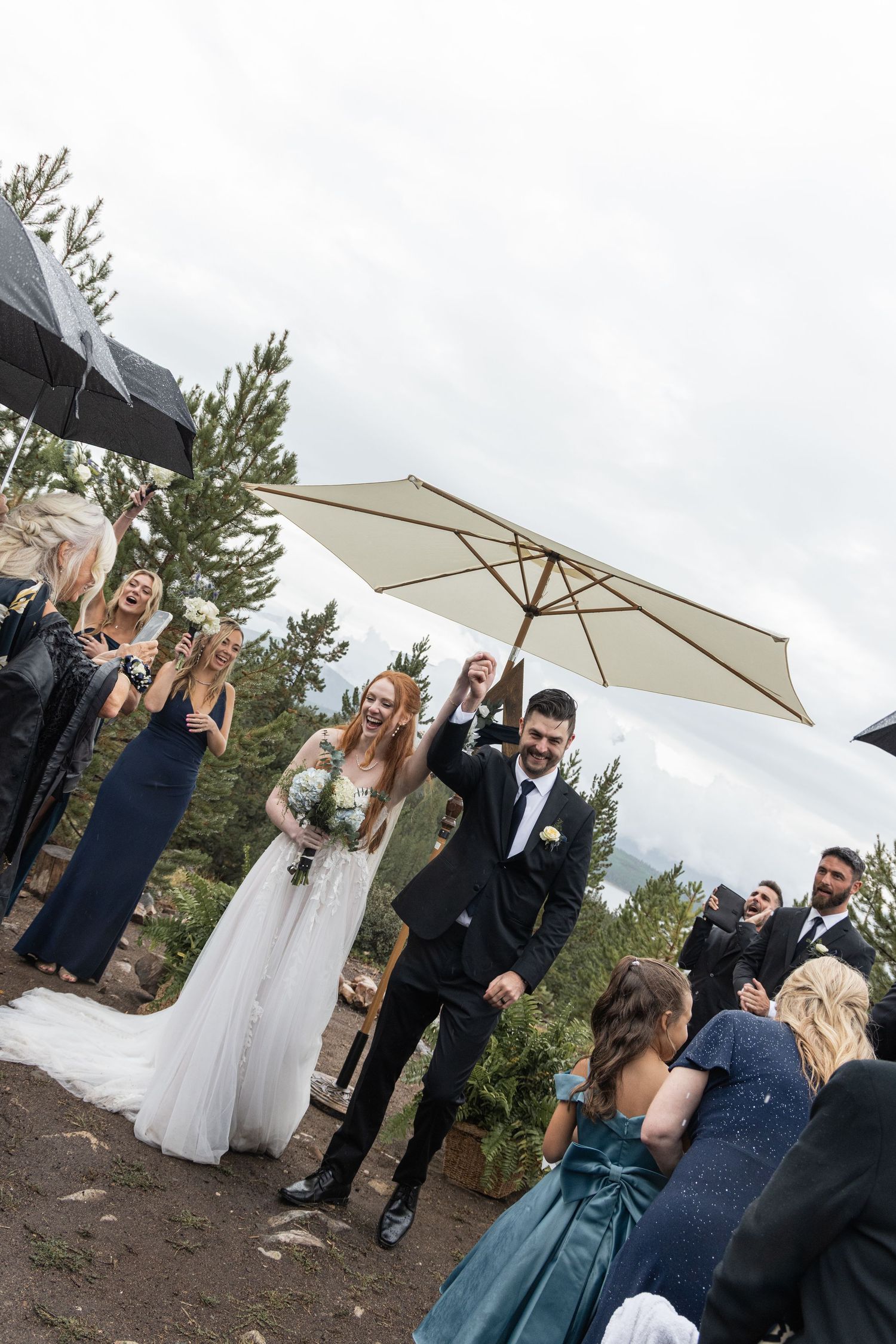 Wedding party celebrates with umbrellas and parasols on a rustic mountain path surrounded by pine trees.