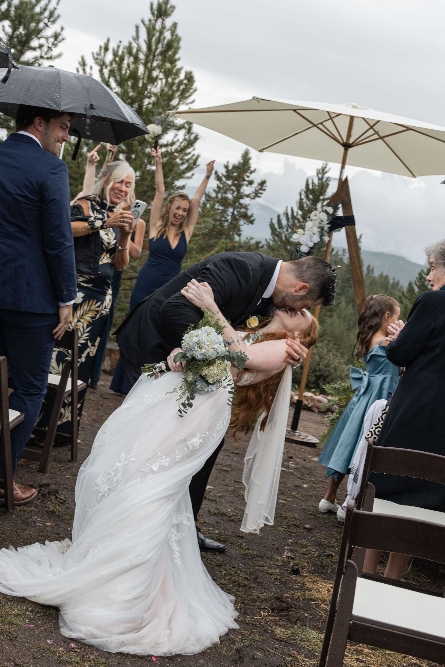 A couple shares a romantic dance at their outdoor mountain wedding reception under white umbrellas and pine trees.