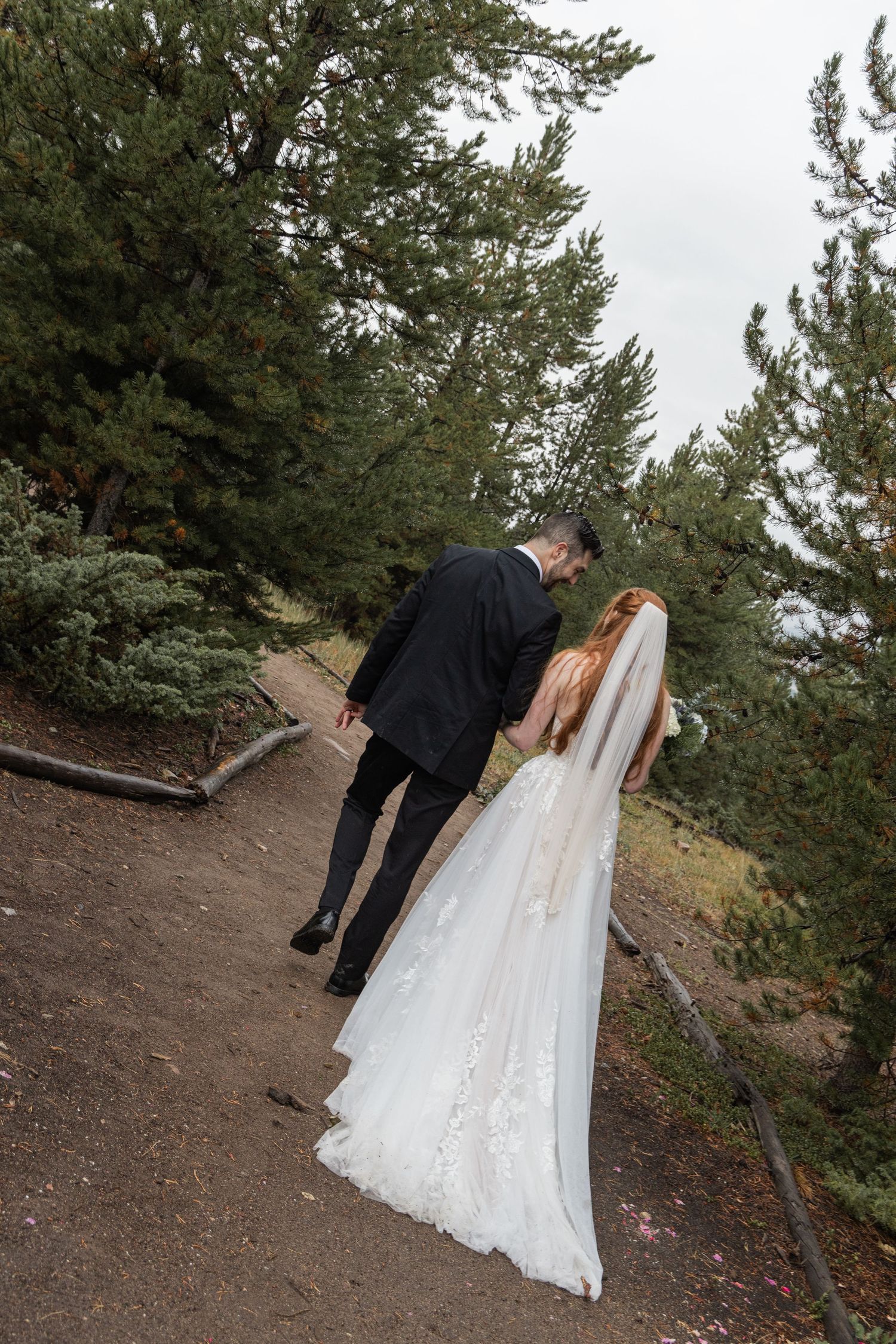 A couple in wedding attire walks along a scenic mountain trail surrounded by evergreen trees.