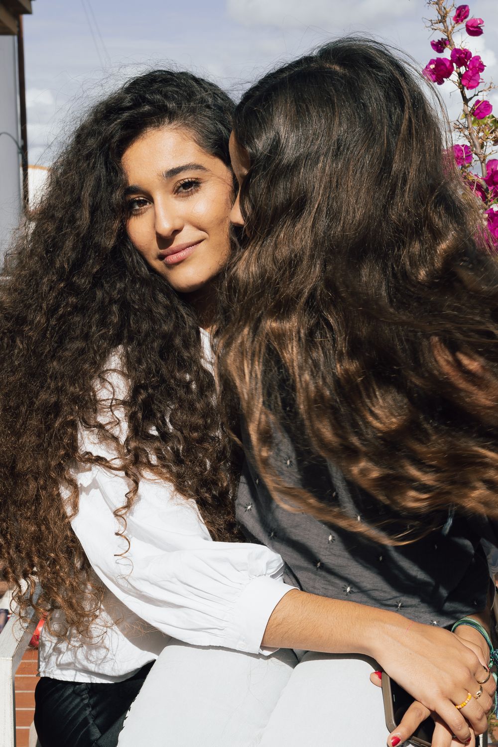 Two people with long curly dark hair sit together near pink flowers outdoors.