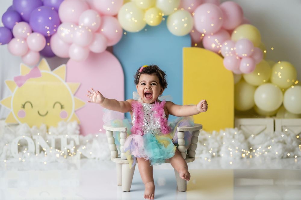 First birthday celebration with pastel balloon arch and baby in colorful outfit.