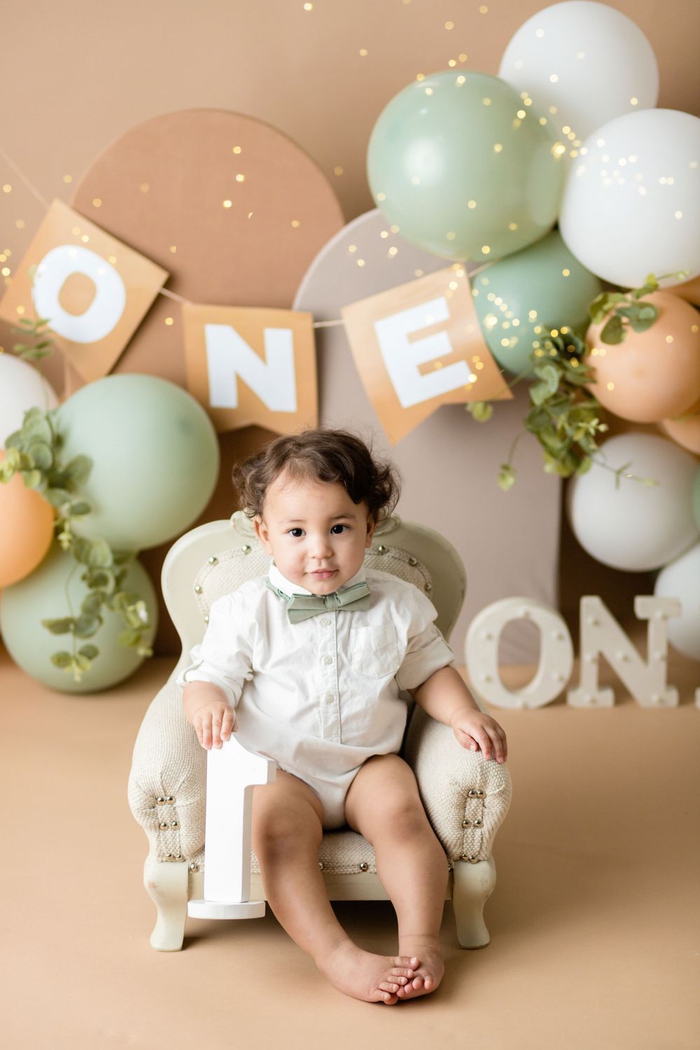 Festive first birthday setup with mint and peach balloons, 'ONE' banner, and cream armchair against neutral backdrop.