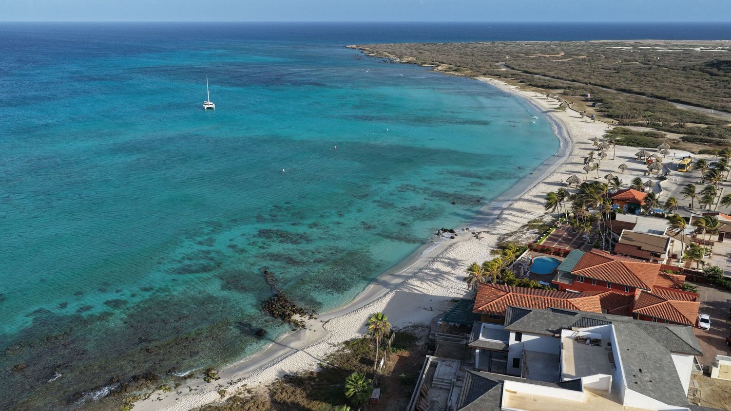 Aerial view of a luxury beachfront resort with turquoise waters, white sand, and beach umbrellas along the coastline.
