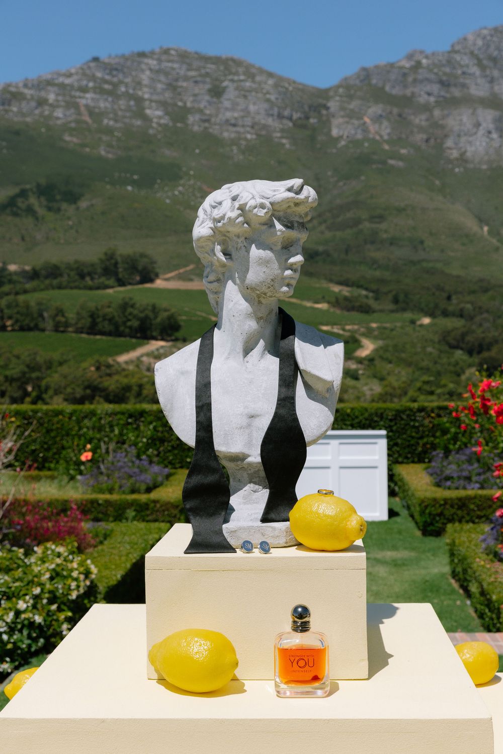 White bust sculpture with lemons and perfume bottle displayed in garden setting with mountains in background.