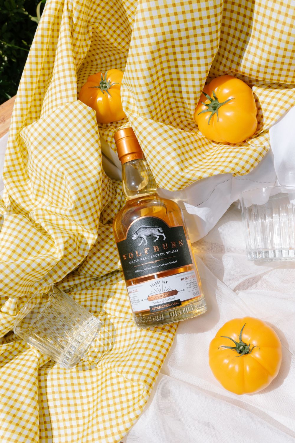 Artfully arranged yellow tomatoes and whiskey bottles on a checkered yellow cloth in natural light.