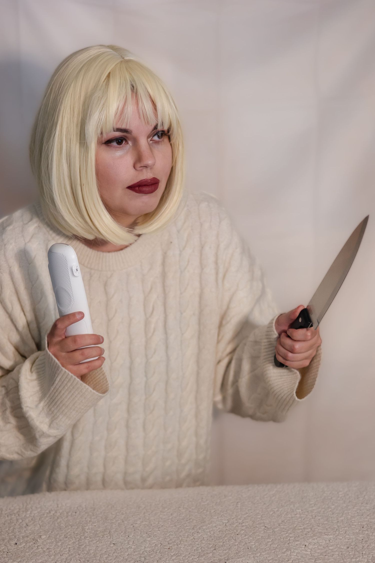 Someone in a cream cable knit sweater holds a knife and remote control with dramatic red lipstick and blonde bob hairstyle.